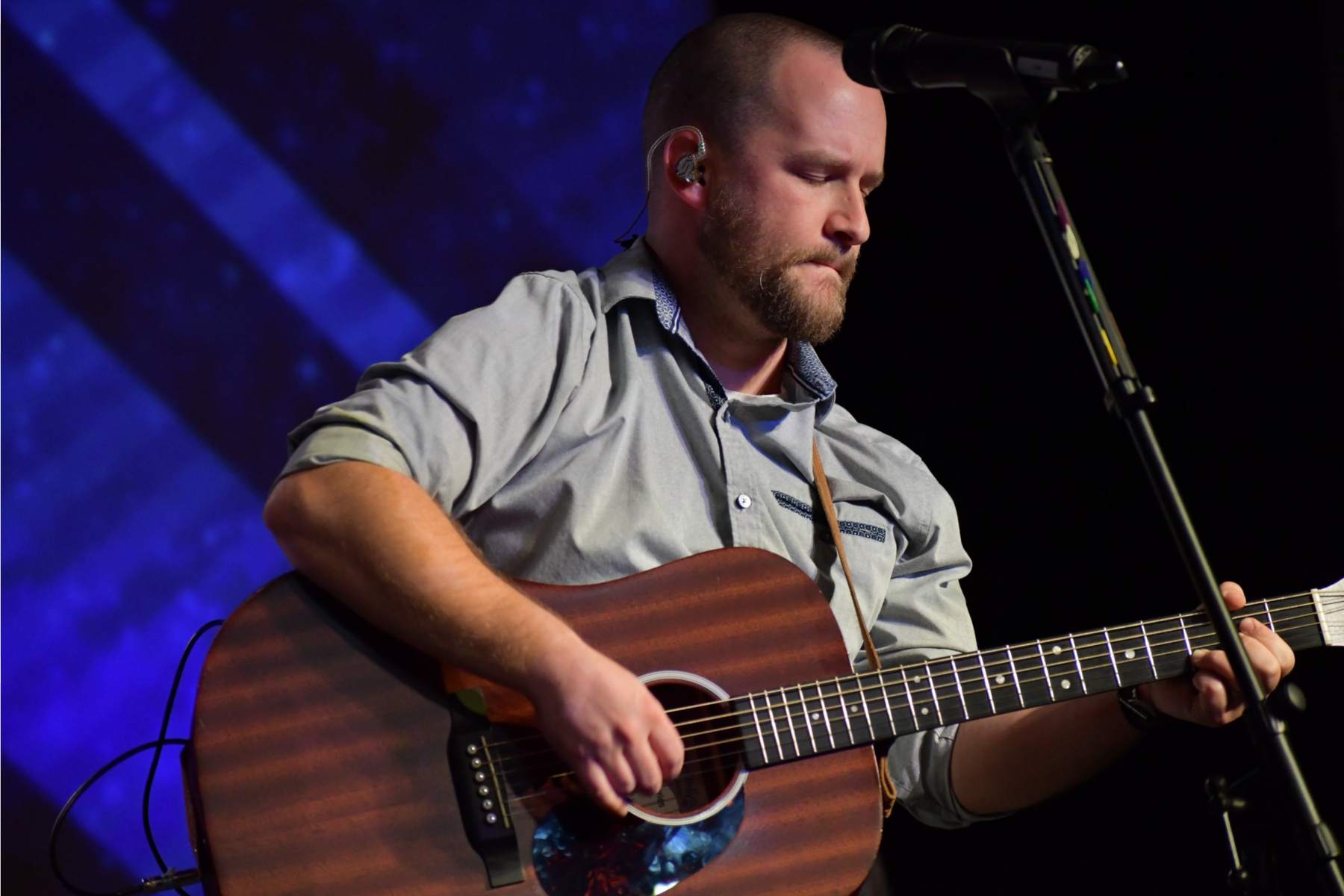 Harry McCarthy playing acoustic guitar on stage under a blue wash, eyes closed, in-ear monitor in, mic stand beside him